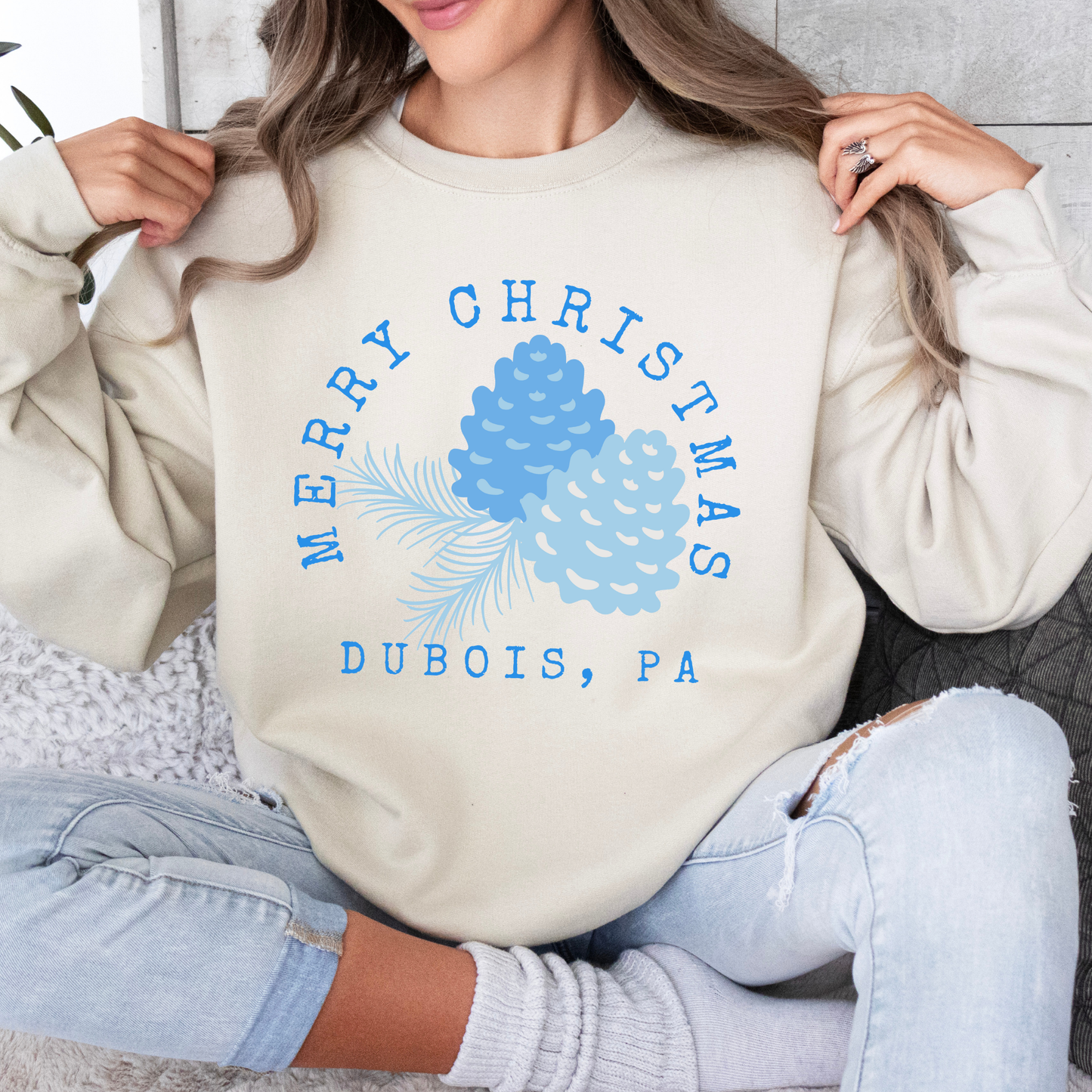 Sand sweatshirt that says Merry Christmas in blue and surrounding blue Christmas pinecones.  below you can customized the sweatshirt with your business name, town and state or both