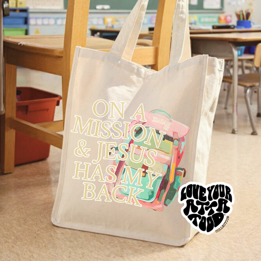 Jumbo canvas tote bag with backpack graphic and “On a Mission and Jesus Has My Back” faith message, statement imagery with painted texture look, Love your ATTA TOOD® brand mark, printed on both sides.