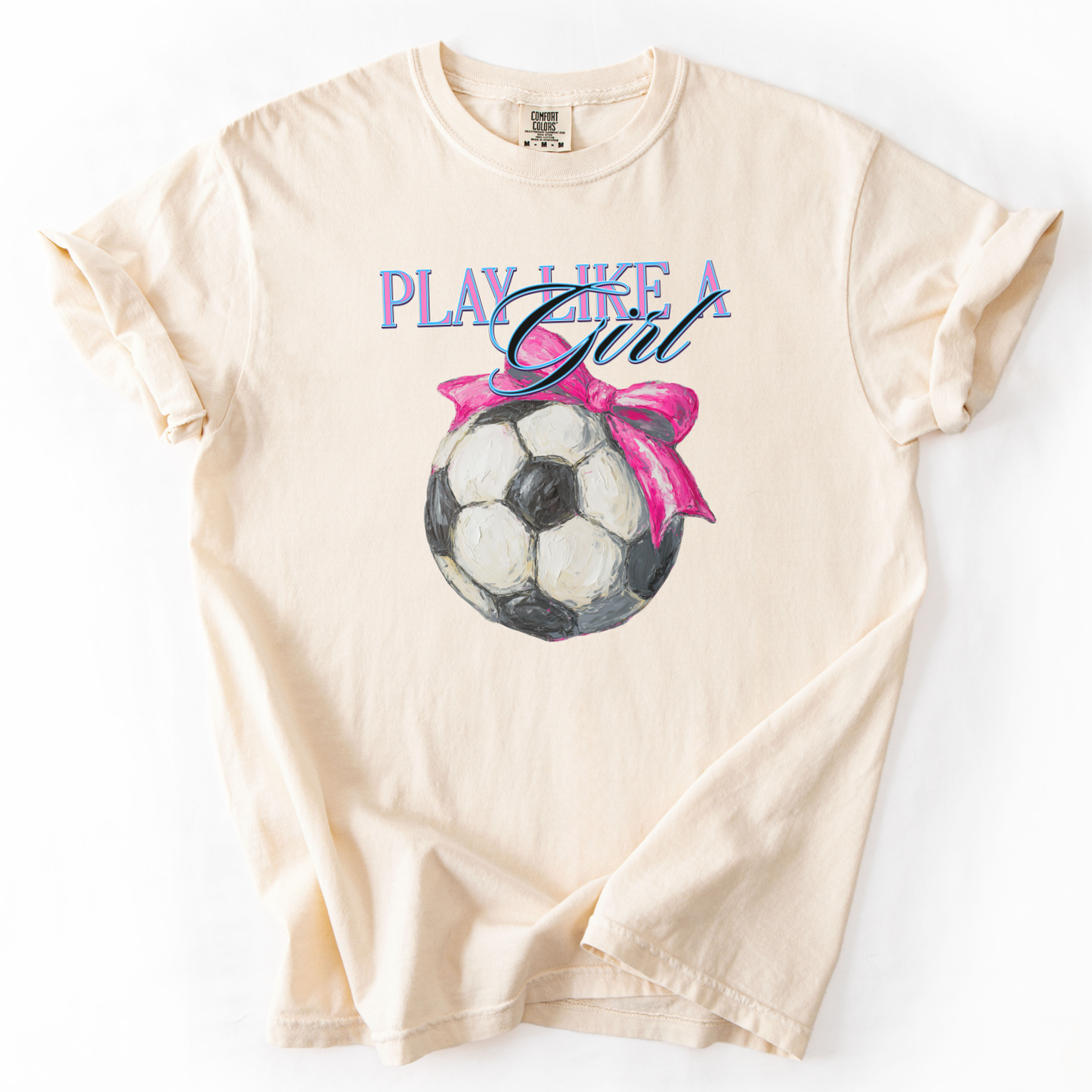 Beige t-shirt with soccer ball and pink bow graphic and 'Play Like a Girl' text on a white background