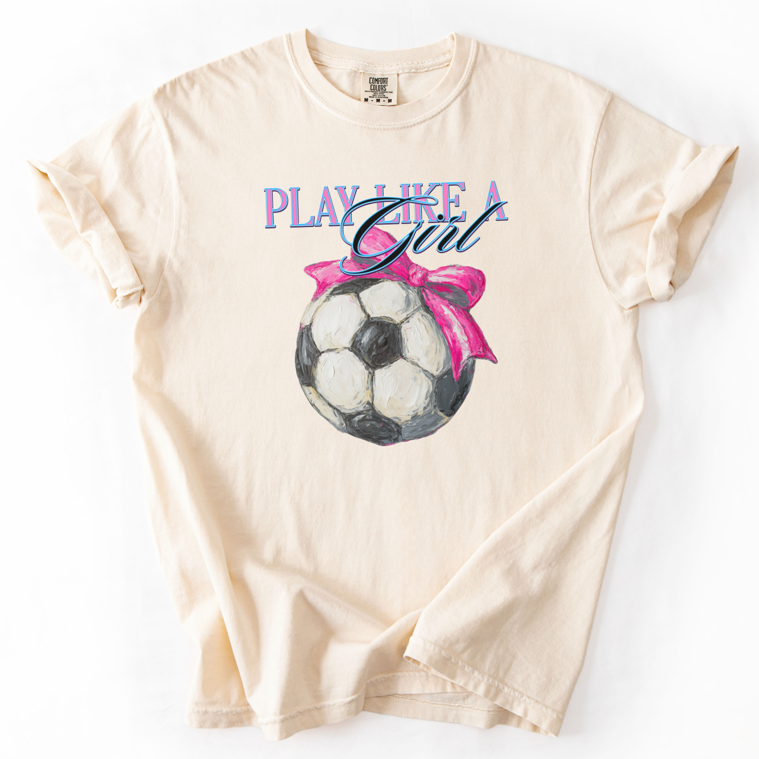 Beige t-shirt with soccer ball and pink bow graphic and 'Play Like a Girl' text on a white background