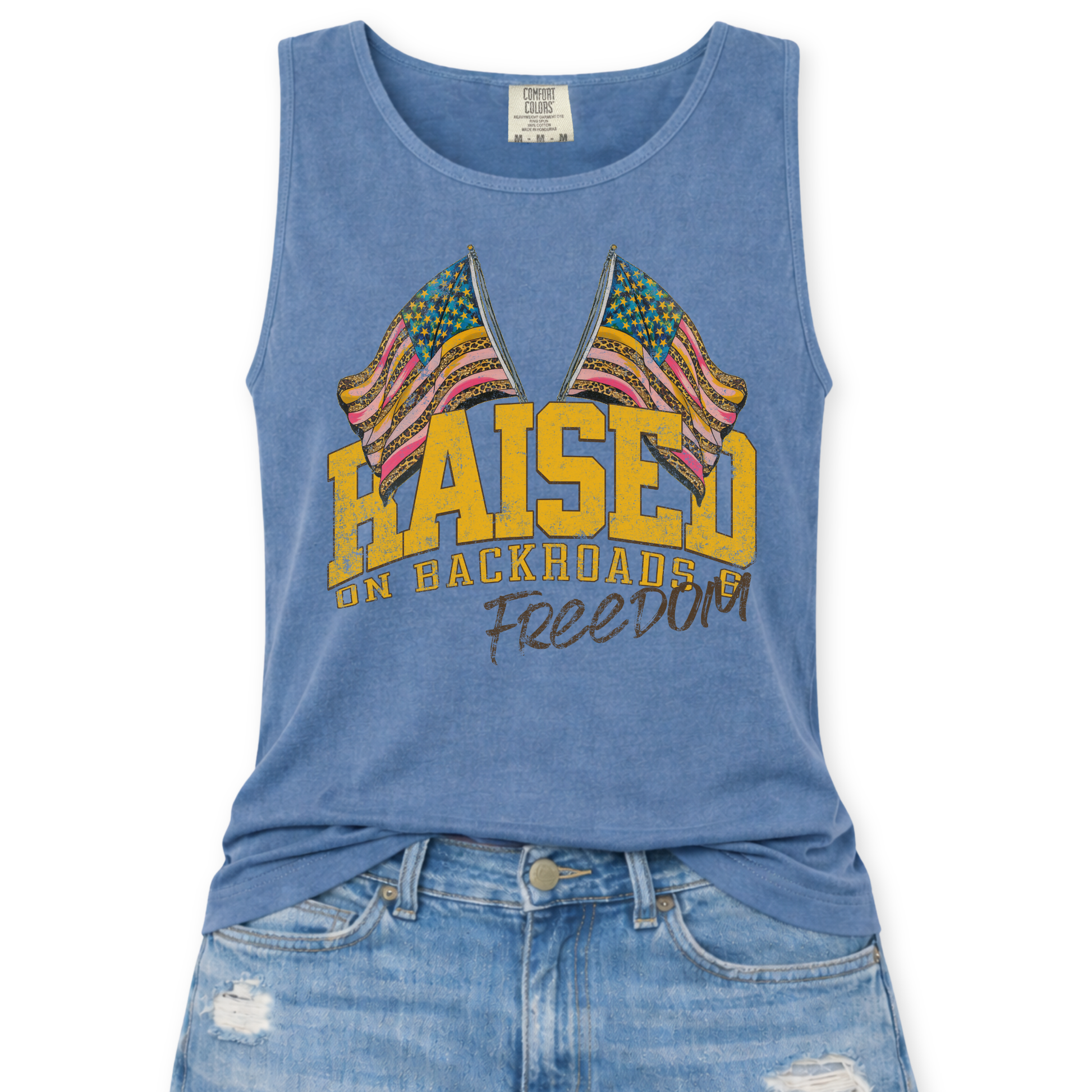 Blue Jean Comfort Colors tank top featuring a distressed patriotic graphic with two crossed American flags above the phrase “Raised on Backroads Freedom” in bold varsity lettering, styled with light wash denim jeans.