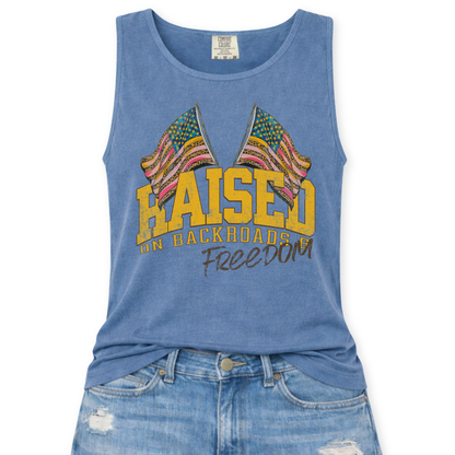 Blue Jean Comfort Colors tank top featuring a distressed patriotic graphic with two crossed American flags above the phrase “Raised on Backroads Freedom” in bold varsity lettering, styled with light wash denim jeans.
