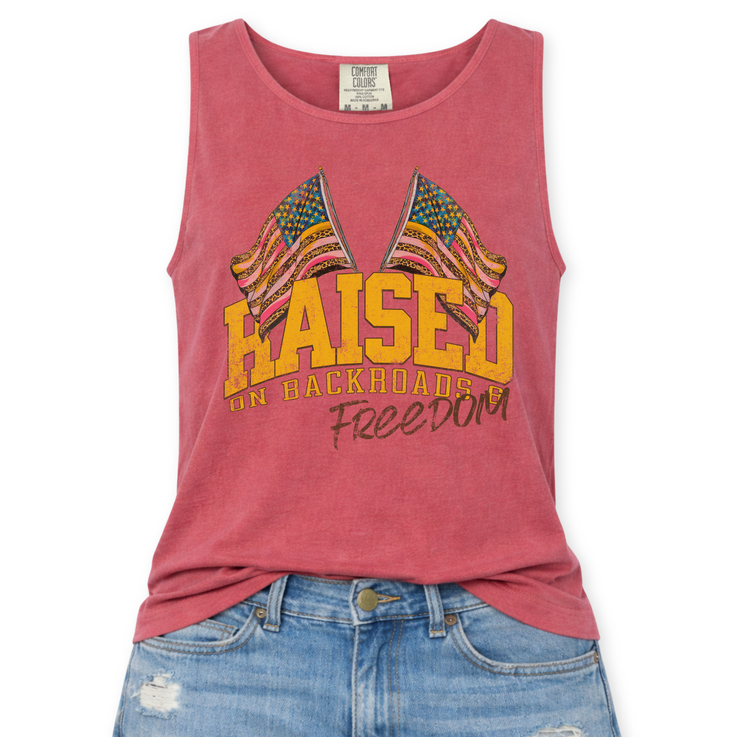 Crimson red Comfort Colors tank top featuring a distressed patriotic graphic with two crossed American flags above the phrase “Raised on Backroads Freedom” in bold varsity lettering, styled with light wash denim jeans.