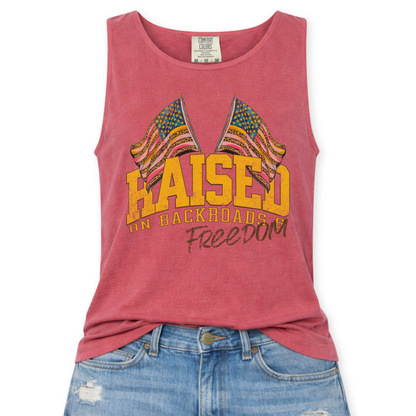 Crimson red Comfort Colors tank top featuring a distressed patriotic graphic with two crossed American flags above the phrase “Raised on Backroads Freedom” in bold varsity lettering, styled with light wash denim jeans.
