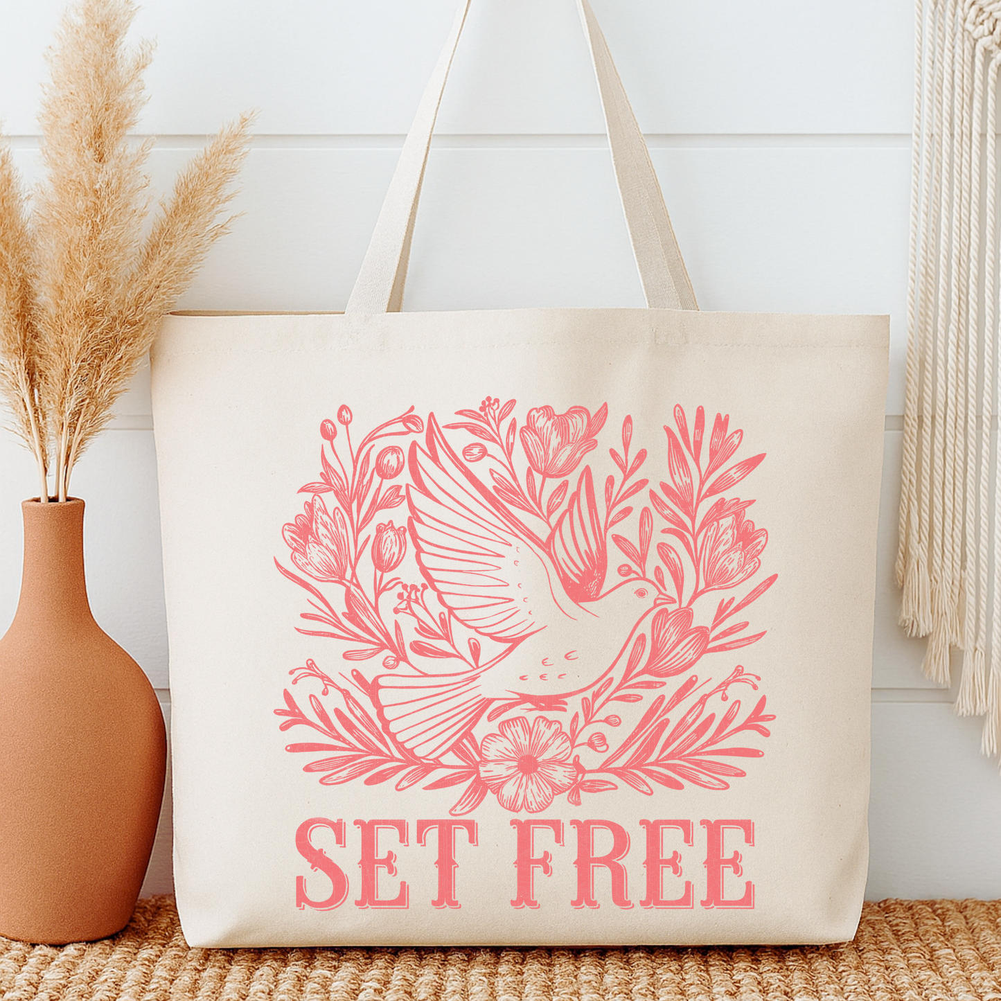 Beige tote bag with pink bird and floral design and 'SET FREE' text, next to a vase with dried plants.