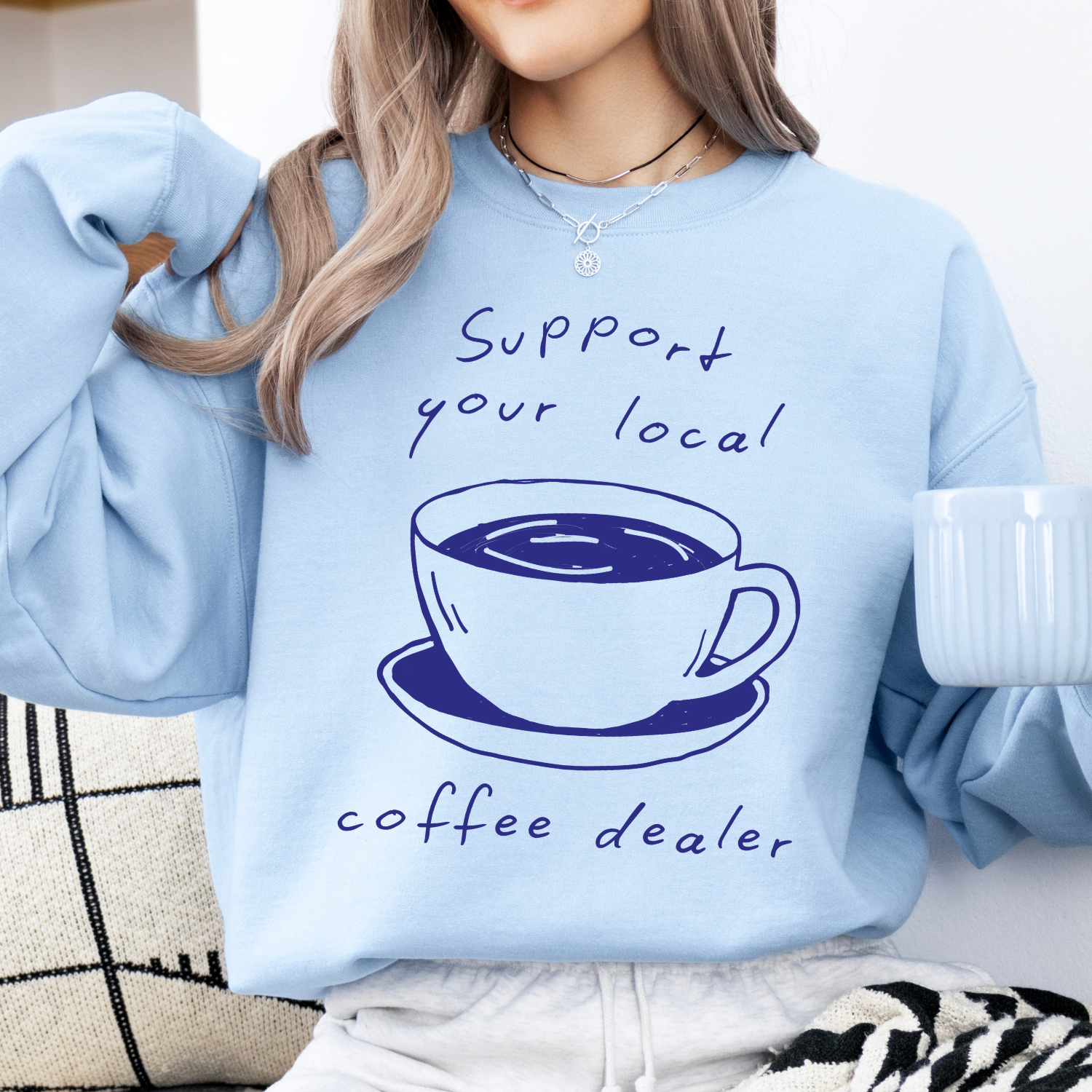 A woman wearing a light blue crewneck sweatshirt with a dark blue graphic of a coffee cup and the text 'Support your local coffee dealer'.