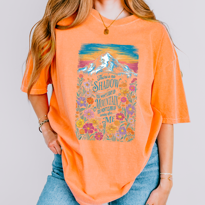 Person wearing an orange t-shirt with a mountain and floral design on a plain background.  There's no shadow you won't light up, Mountain you won't climb up coming after me