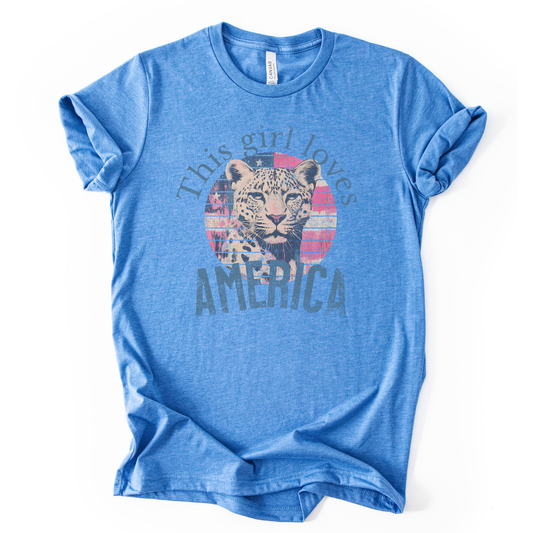 Patriotic Americana graphic on a blue t-shirt. The design features an illustrated leopard portrait centered over a distressed American flag circle, paired with the phrase “This Girl Loves America” in bold, vintage-style lettering. The worn texture and soft color palette create a modern patriotic look that blends animal imagery with classic Fourth of July appeal.