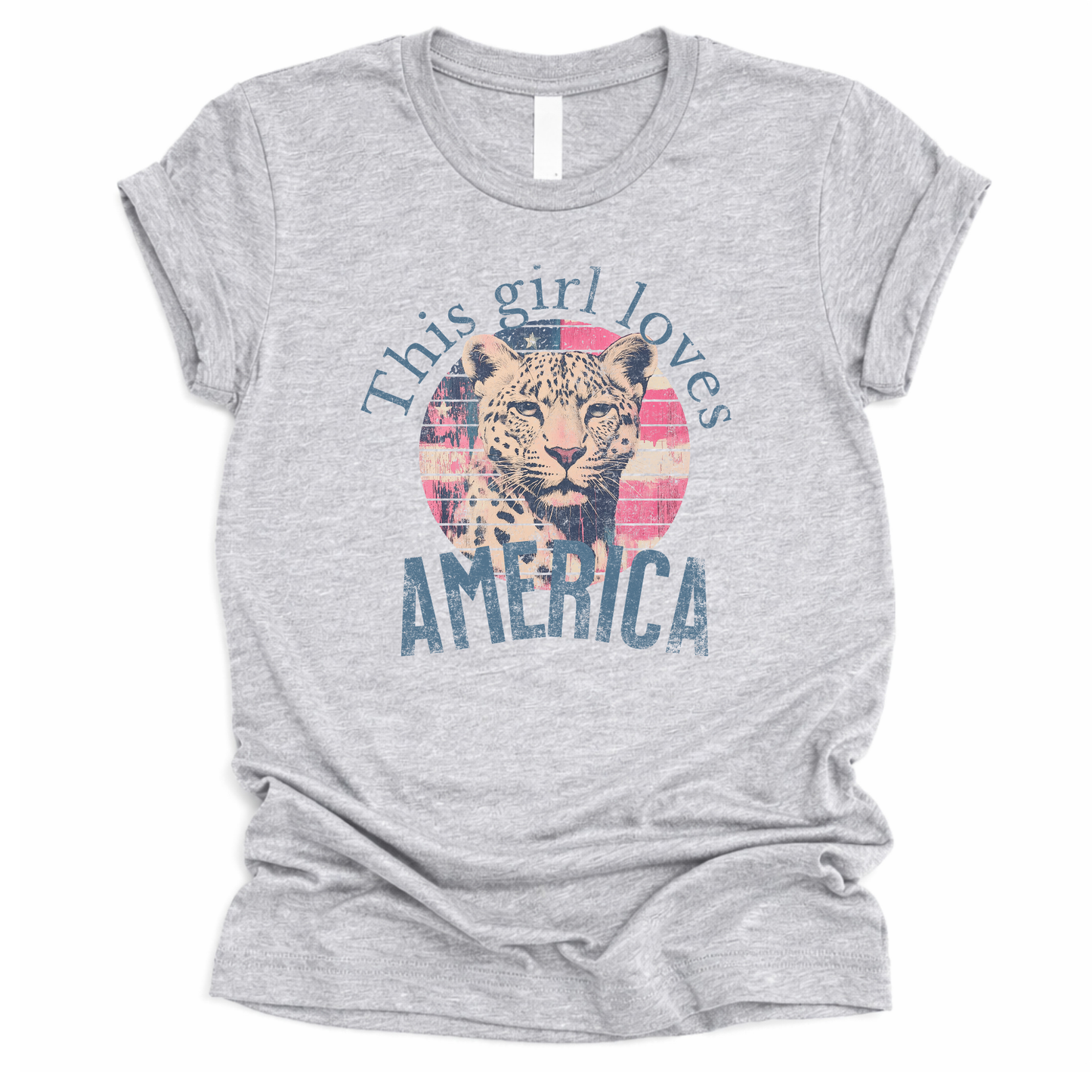 Patriotic Americana graphic on a gray t-shirt. The design features an illustrated leopard portrait centered over a distressed American flag circle, paired with the phrase “This Girl Loves America” in bold, vintage-style lettering. The worn texture and soft color palette create a modern patriotic look that blends animal imagery with classic Fourth of July appeal.