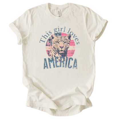Patriotic Americana graphic on a beige t-shirt. The design features an illustrated leopard portrait centered over a distressed American flag circle, paired with the phrase “This Girl Loves America” in bold, vintage-style lettering. The worn texture and soft color palette create a modern patriotic look that blends animal imagery with classic Fourth of July appeal.