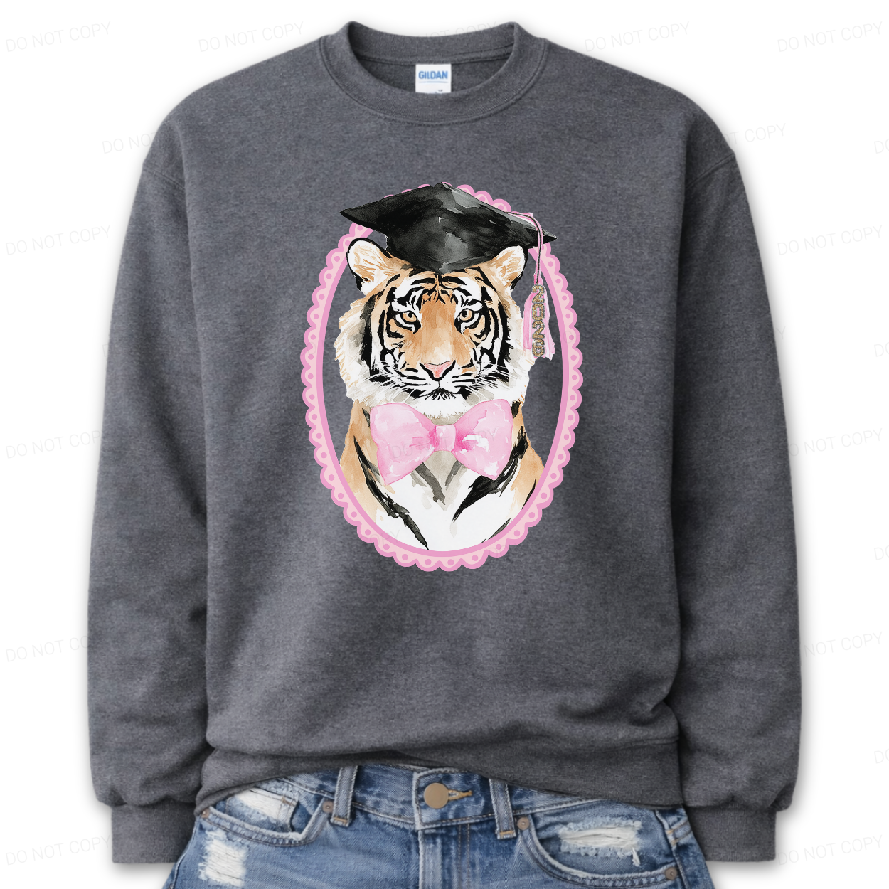Graduation tiger graphic on a classic crewneck sweatshirt. The design features a detailed tiger face illustration framed in a soft pink oval, wearing a black graduation cap with tassel and finished with a pink bow at the neckline. The mascot style artwork with preppy pink accents creates a standout graduation season look that works for both high school seniors and college graduates.
