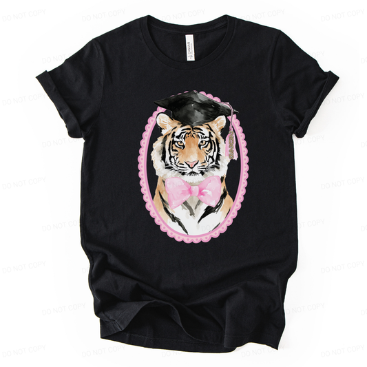 Graduation 2026 mascot themed graphic t shirt. The design features a detailed tiger portrait wearing a black graduation cap with a 2026 tassel, accented with a soft pink bow and framed in a decorative oval border. The combination of mascot styling and feminine pink accents creates a strong senior year statement with clear Class of 2026 visibility.