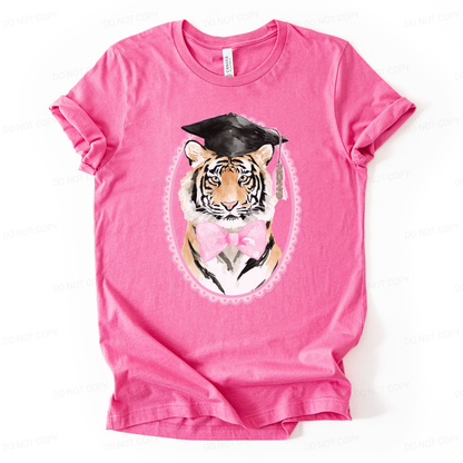 Graduation 2026 mascot themed graphic t shirt. The design features a detailed tiger portrait wearing a black graduation cap with a 2026 tassel, accented with a soft pink bow and framed in a decorative oval border. The combination of mascot styling and feminine pink accents creates a strong senior year statement with clear Class of 2026 visibility.