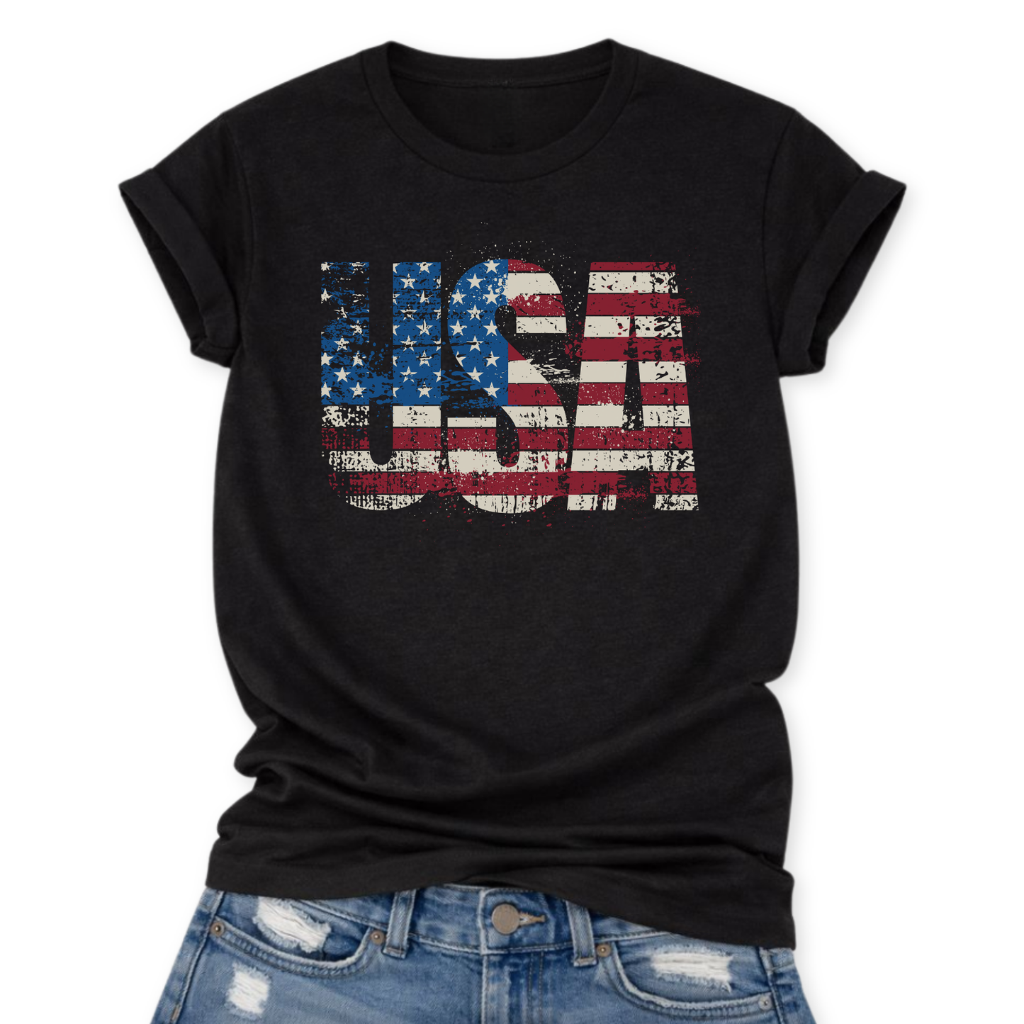 Black Bella Canvas t-shirt featuring large distressed “USA” block letters filled with an American flag print in red, white, and blue, shown in a flat lay with a slight front tuck into distressed denim.