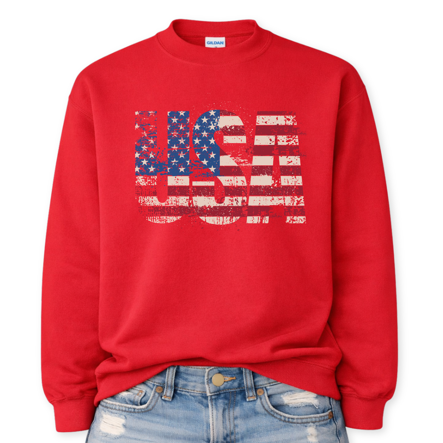 Gildan red crewneck sweatshirt mockup with a front tuck into distressed denim, featuring oversized “USA” block letters filled with a distressed American flag stars-and-stripes graphic.