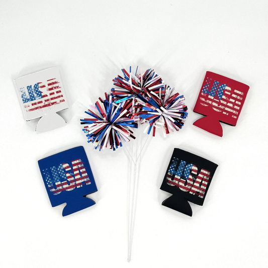 Assorted collapsible can coolers in white, red, royal blue, and black featuring a distressed American flag “USA” graphic. Displayed with red, white, and blue metallic pom poms on a white background.