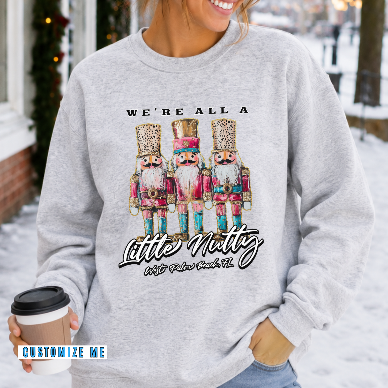A woman wearing a light heather gray sweatshirt with a graphic of three nutcracker children and the text 'WE'RE ALL A Little Nutty With Faber Buck Co.' while holding a coffee cup in a snowy outdoor setting.