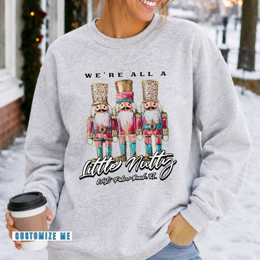A woman wearing a light heather gray sweatshirt with a graphic of three nutcracker children and the text 'WE'RE ALL A Little Nutty With Faber Buck Co.' while holding a coffee cup in a snowy outdoor setting.