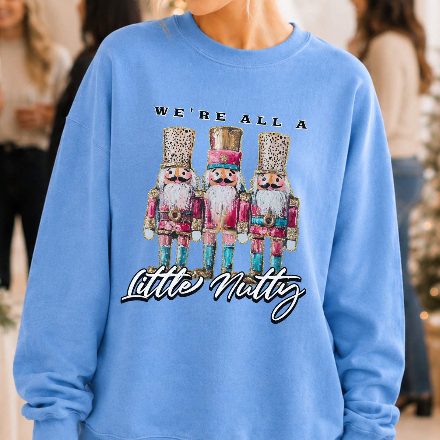 A person wearing a light blue sweatshirt with a graphic of three nutcracker figures and the text 'WE'RE ALL A Little Natty' printed on the front, with a softly blurred indoor background.