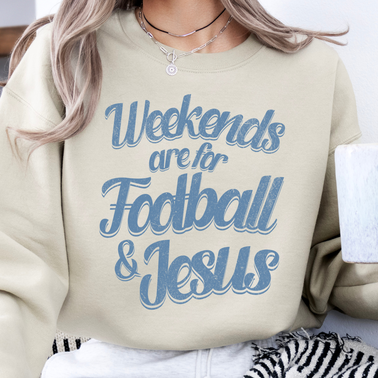 A woman wearing a beige sweatshirt with the text 'Weekends are for FOOTBALL & JESUS' printed on the front, holding a coffee cup, with a zebra-patterned blanket and a pendant visible in the background.