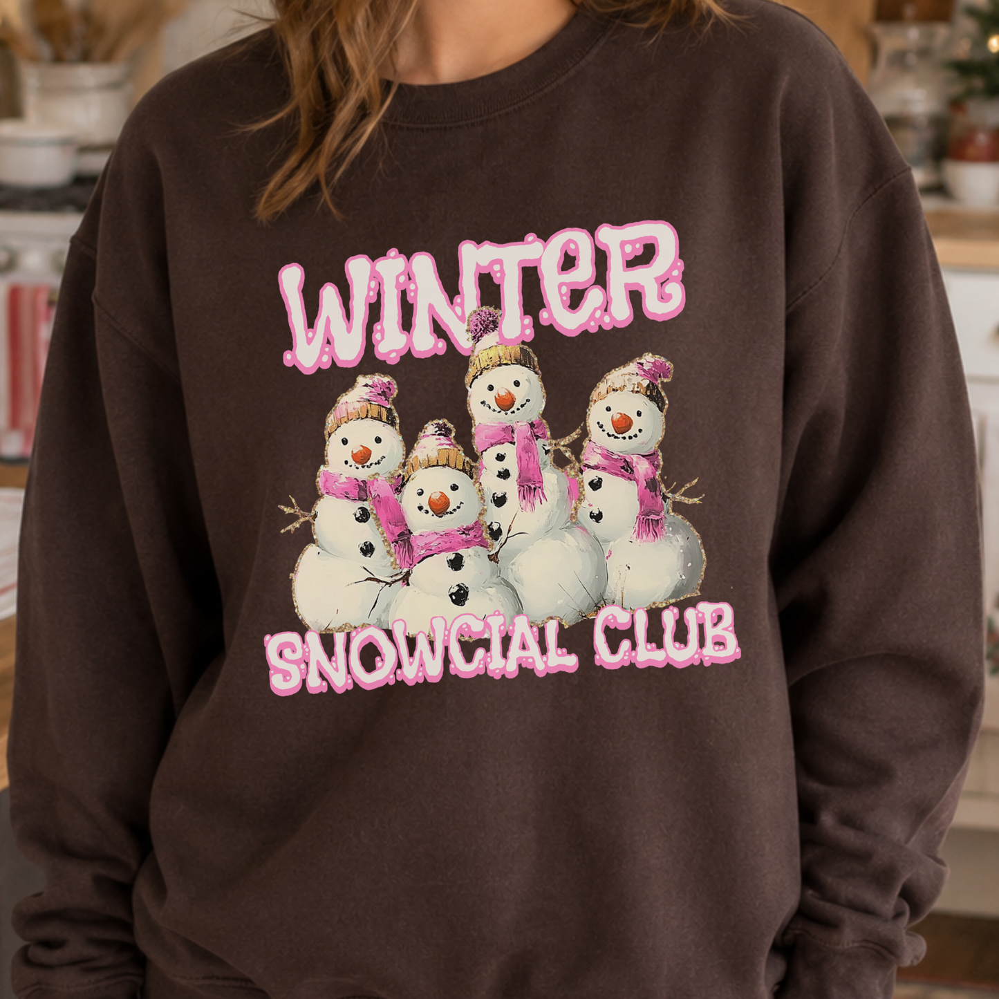 A person wearing a dark brown crewneck sweatshirt with a graphic print of six snowmen wearing pink scarves and hats under the word 'WINTER' and above 'SNOWCIAL CLUB'.