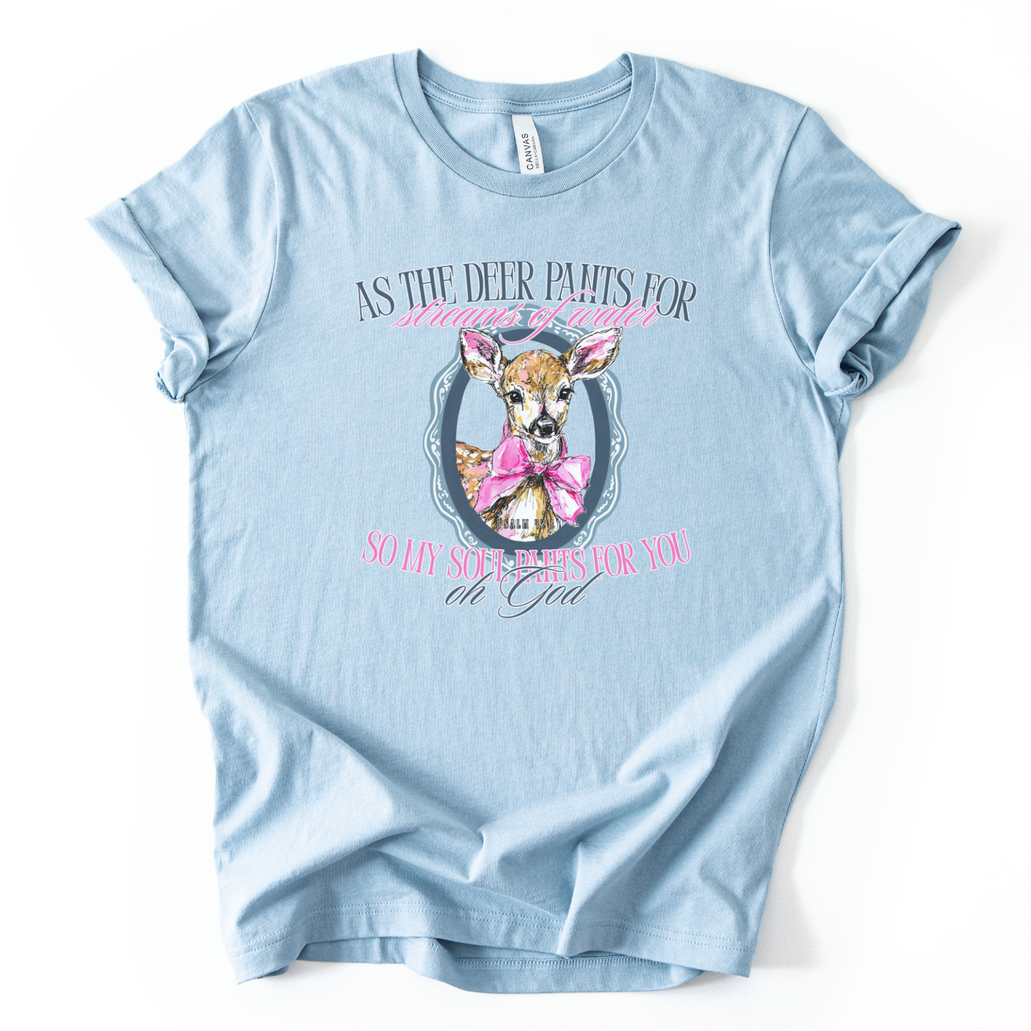 The graphic features a illustrated deer portrait centered inside a decorative oval frame, styled with a large pink bow around the deer’s neck. The design is paired with the scripture text “As the deer pants for streams of water, so my soul pants for You, oh God” (Psalm 42:1) in soft, feminine lettering.  Image is on a baby blue heather Bella Canvas Tee
