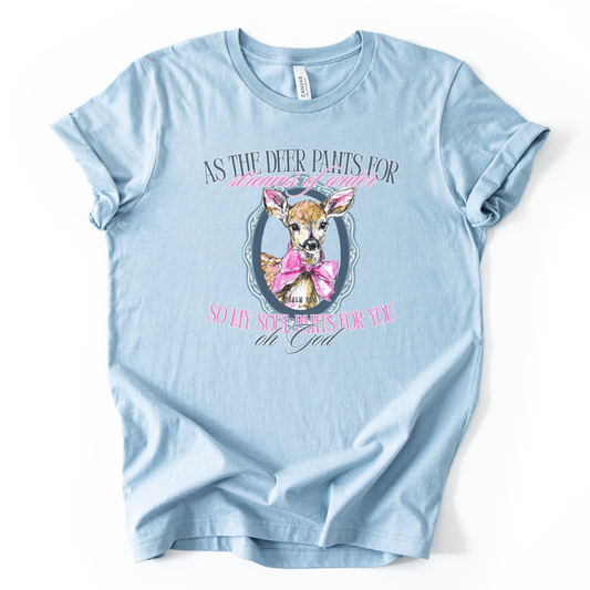 The graphic features a illustrated deer portrait centered inside a decorative oval frame, styled with a large pink bow around the deer’s neck. The design is paired with the scripture text “As the deer pants for streams of water, so my soul pants for You, oh God” (Psalm 42:1) in soft, feminine lettering.  Image is on a baby blue heather Bella Canvas Tee