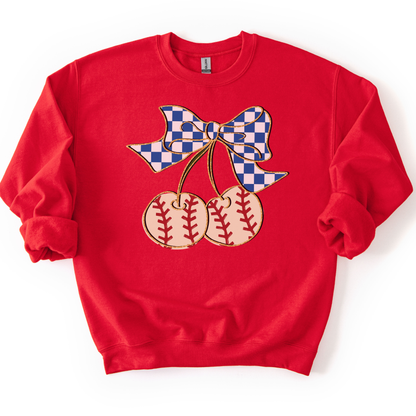 Red crewneck sweatshirt featuring a blue gingham bow tied to two cherry-shaped baseballs with red stitching, styled flat with sleeves scrunched