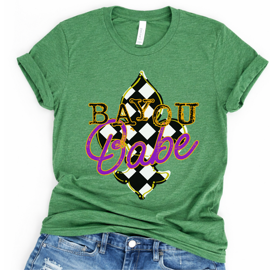 Flat lay of a Bella Canvas unisex graphic t-shirt featuring a “Bayou Babe” design with bold lettering over a black and white harlequin pattern, styled casually with denim for a southern-inspired, Mardi Gras-ready look.