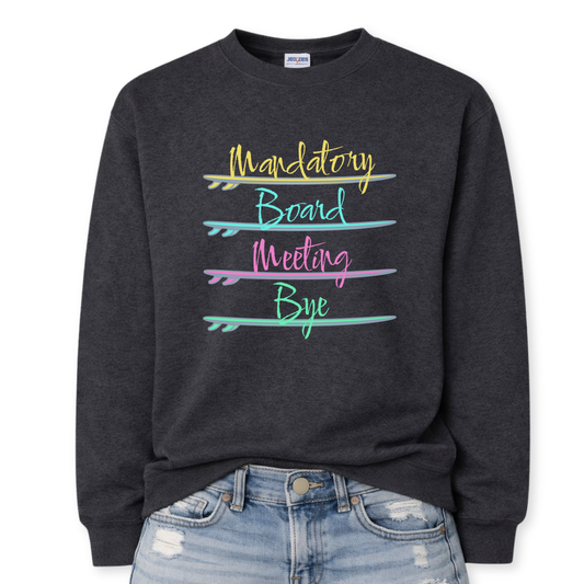 Beach humor graphic on a classic dark heather gray crewneck sweatshirt. The design features stacked lettering that reads Mandatory Board Meeting Bye, with each word separated by illustrated surfboards that act as graphic dividers. The colorful surfboards create a playful coastal layout that reinforces the beach lifestyle theme.