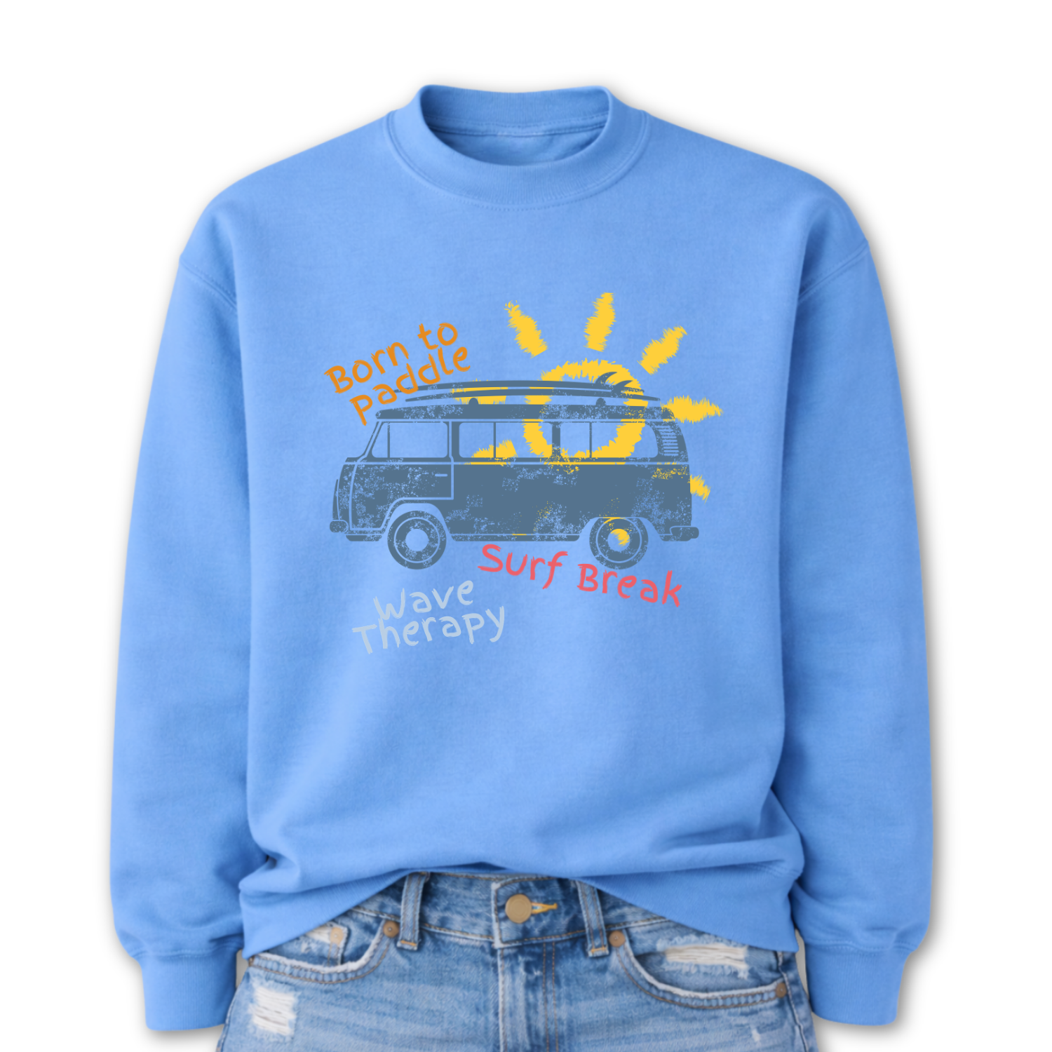  Beach surf lifestyle graphic on a classic blue crewneck sweatshirt. The design features a retro surf van with a surfboard mounted on the roof, set in front of a stylized sun graphic. The phrases Born To Paddle, Surf Break, and Wave Therapy surround the van illustration, creating a layered beach adventure layout inspired by surf culture and coastal travel.
