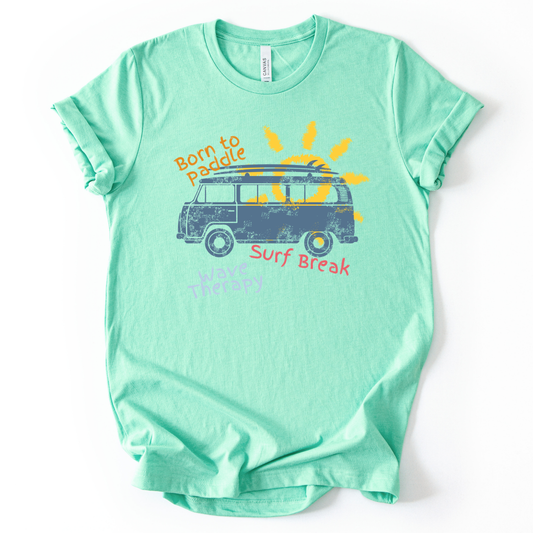 Summer beach graphic on a soft unisex mint green graphic tee. The design features a vintage surf van illustration with a surfboard on top and a bright sun rising behind the vehicle. Playful coastal phrases including Born to Paddle, Surf Break, and Wave Therapy surround the artwork, creating a relaxed surf and beach lifestyle graphic that captures the feeling of ocean days and summer travel.