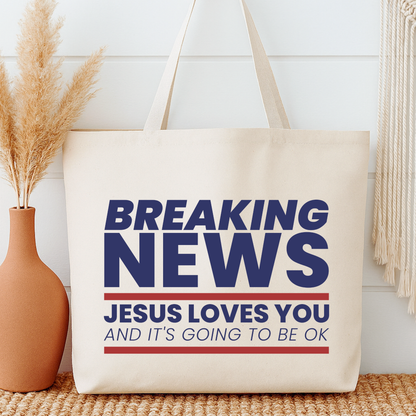 Beige tote bag with 'Breaking News: Jesus Loves You and It's Going to Be Ok' text, next to a vase with dried plants.