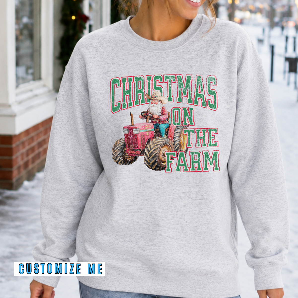 A woman wearing a light heather gray sweatshirt with a 'Christmas on the Farm' graphic featuring a red tractor and green lettering, set against a snowy outdoor background.