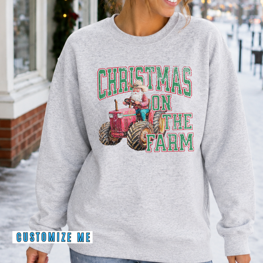 A woman wearing a light heather gray sweatshirt with a 'Christmas on the Farm' graphic featuring a red tractor and green lettering, set against a snowy outdoor background.