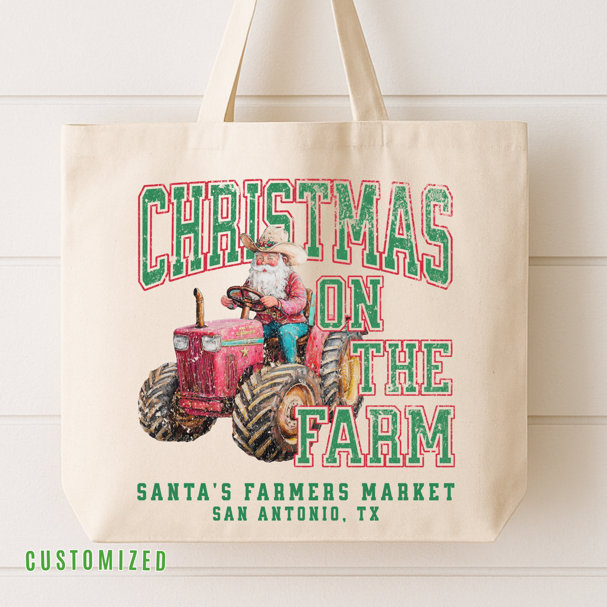 A cream canvas tote bag with a festive 'Christmas on the Farm' graphic featuring Santa driving a red tractor, with 'Santa's Farmers Market San Antonio, TX' printed below. The bag is shown hanging against a white wood-paneled wall.