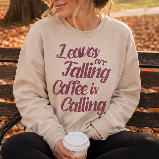 Person wearing a beige sweatshirt with 'Leaves are falling, Coffee is calling' text, sitting on a bench outdoors.