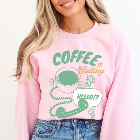 A light pink crewneck sweatshirt featuring a graphic print with the text 'COFFEE is Bailing' in green and orange, accompanied by a green coffee cup, coiled phone cord, green phone receiver, and a speech bubble that says 'HELLO!?' on a white background.