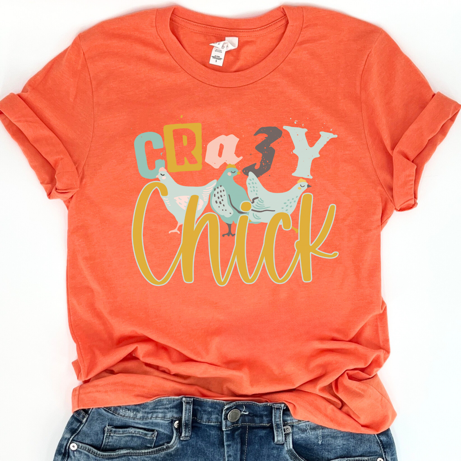 Flat lay of a Bella Canvas unisex graphic t-shirt featuring the phrase “Crazy Chick” with illustrated chickens in playful typography, styled casually with denim for a fun, farm-inspired everyday look.