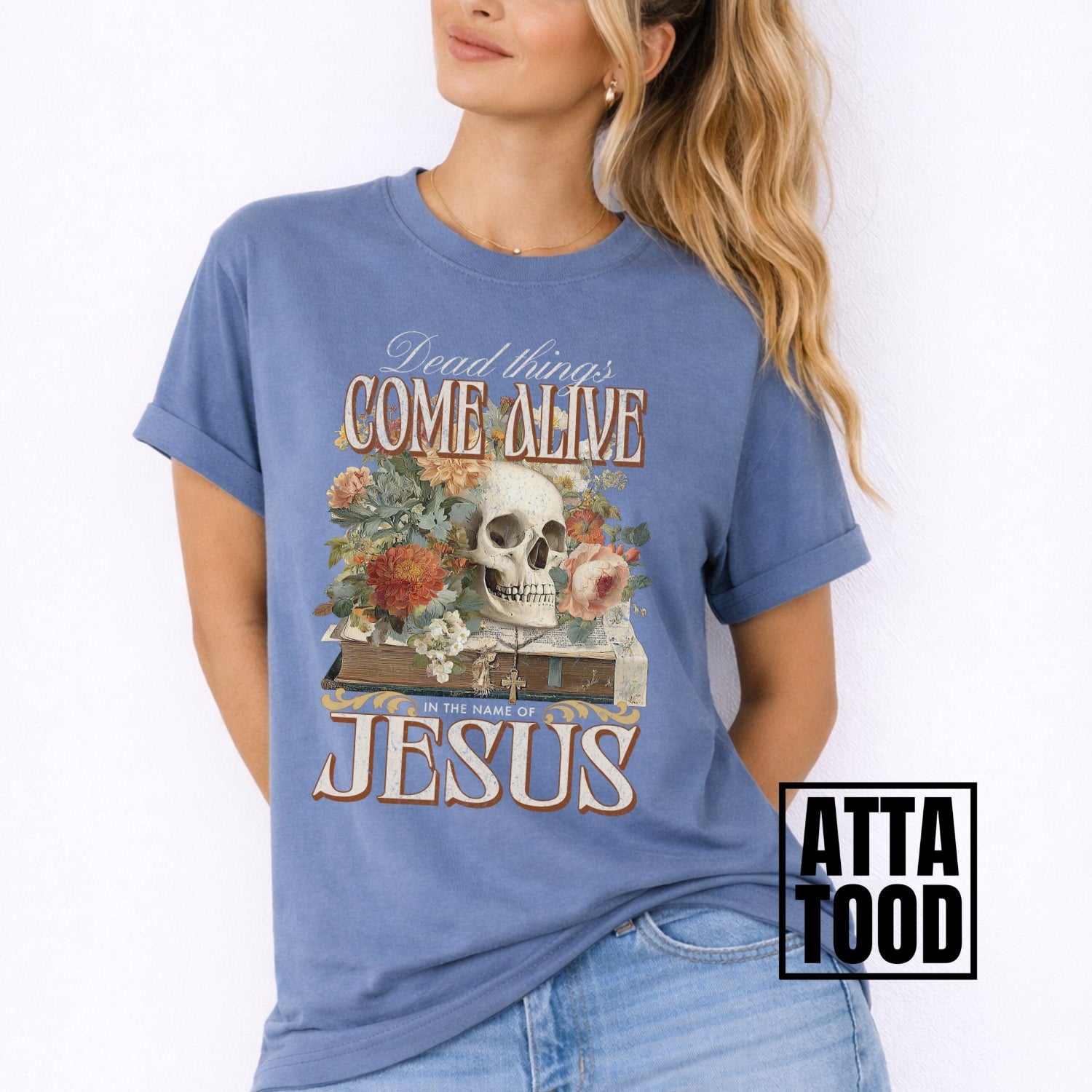 Dead things come alive in the name of Jesus with an oversized graphic of a bible, skeleton head and flowers on a comfort colors blue tee