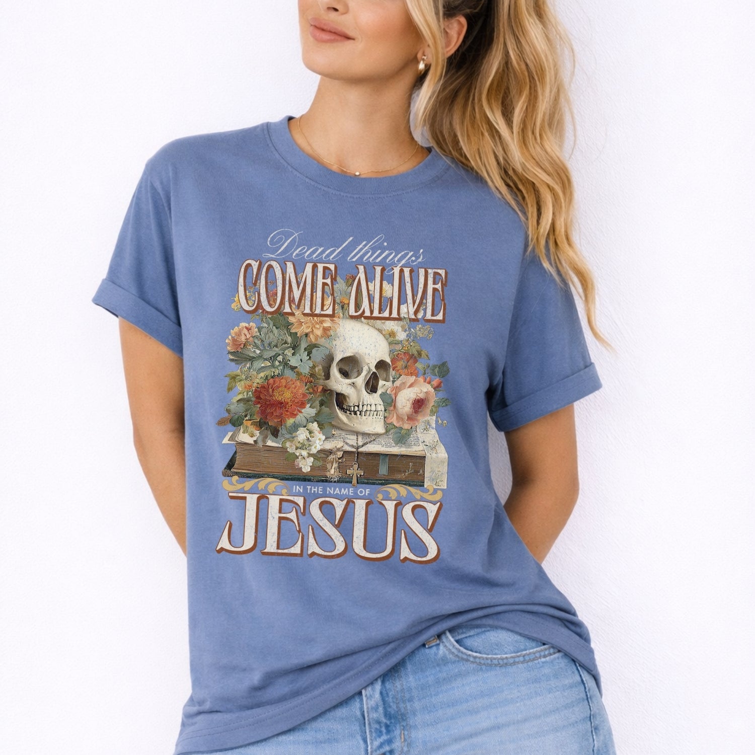 Dead things come alive in the name of Jesus with an oversized graphic of a bible, skeleton head and flowers on a comfort colors Blue Jean tee