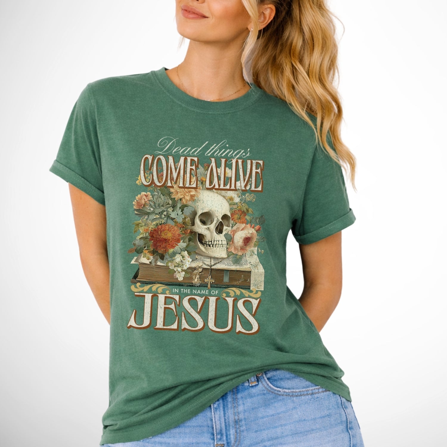 Dead things come alive in the name of Jesus with an oversized graphic of a bible, skeleton head and flowers on a comfort colors light green tee