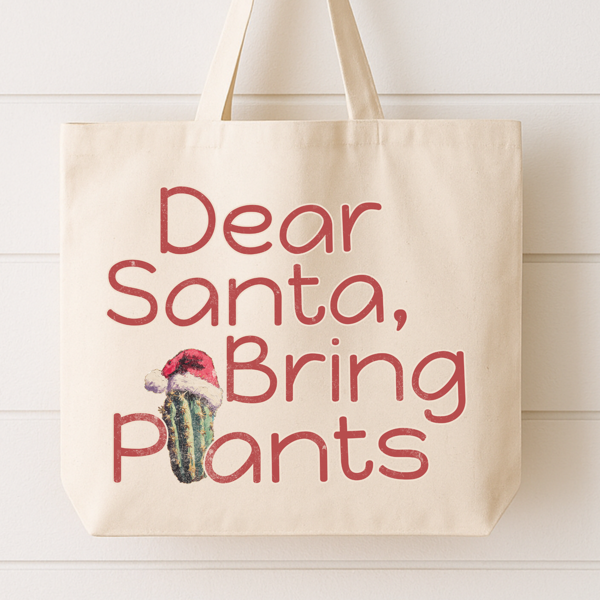 A cream canvas tote bag with the phrase 'Dear Santa, Bring Plants' printed in a dusty red font, featuring a small cactus wearing a Santa hat graphic, hanging against a white wood paneled wall.
