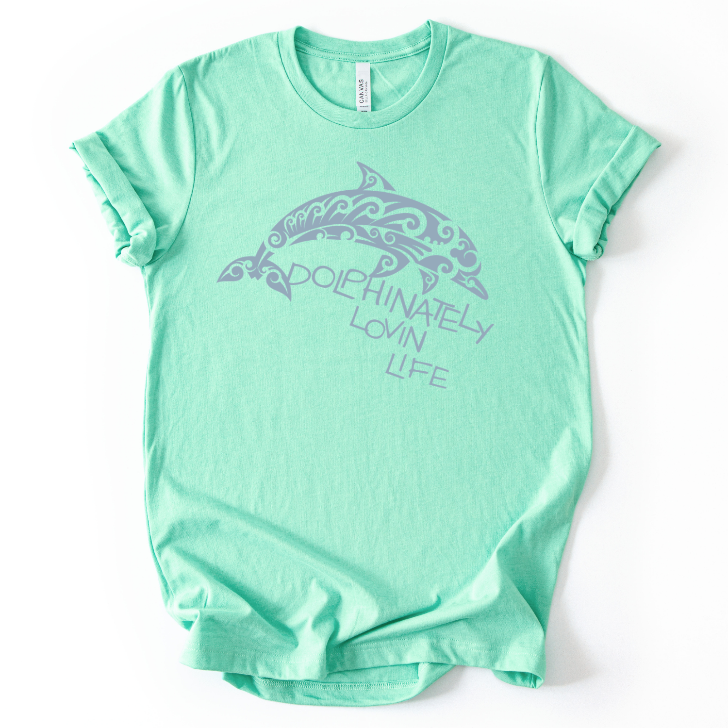 Coastal ocean graphic on a soft unisex heather mint graphic tee. The design features a large dolphin illustration filled with decorative swirl and wave inspired pattern details that give the sea life artwork a textured ocean aesthetic. The phrase Dolphinately Lovin Life appears integrated with the graphic, creating a playful ocean themed message that pairs with the curved shape of the dolphin.