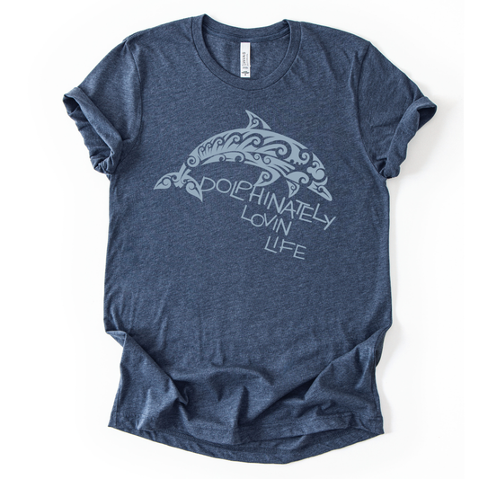 Coastal ocean graphic on a soft unisex heather navy graphic tee. The design features a large dolphin illustration filled with decorative swirl and wave inspired pattern details that give the sea life artwork a textured ocean aesthetic. The phrase Dolphinately Lovin Life appears integrated with the graphic, creating a playful ocean themed message that pairs with the curved shape of the dolphin.