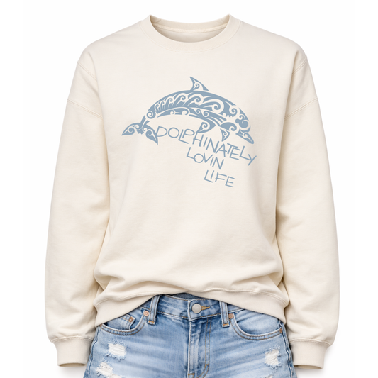 Coastal ocean graphic on a classic ivory crewneck sweatshirt. The design features a large dolphin illustration filled with decorative swirl and wave inspired pattern details that give the sea life artwork a textured ocean aesthetic. The phrase Dolphinately Lovin Life is integrated into the graphic beneath the dolphin.