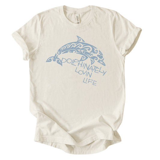 Coastal ocean graphic on a soft unisex heather oatmeal graphic tee. The design features a large dolphin illustration filled with decorative swirl and wave inspired pattern details that give the sea life artwork a textured ocean aesthetic. The phrase Dolphinately Lovin Life appears integrated with the graphic, creating a playful ocean themed message that pairs with the curved shape of the dolphin.