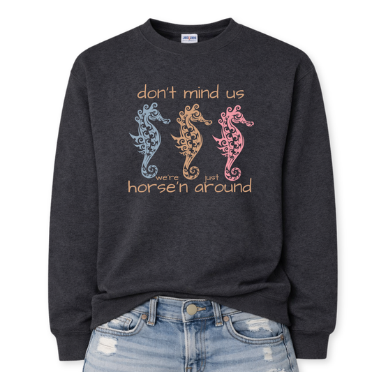 Bold summer coastal graphic on a classic black heather crewneck sweatshirt. The design features three ornamental seahorses in soft coastal colors with the wording don't mind us were just horse'n around. 