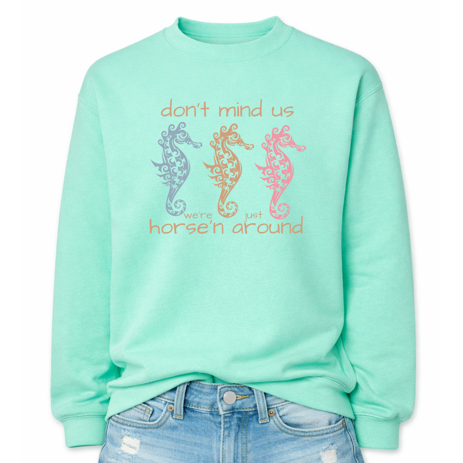 Bold summer coastal graphic on a classic mint green crewneck sweatshirt. The design features three ornamental seahorses in soft coastal colors with the wording don't mind us were just horse'n around. 