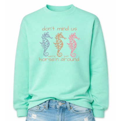Bold summer coastal graphic on a classic mint green crewneck sweatshirt. The design features three ornamental seahorses in soft coastal colors with the wording don't mind us were just horse'n around. 